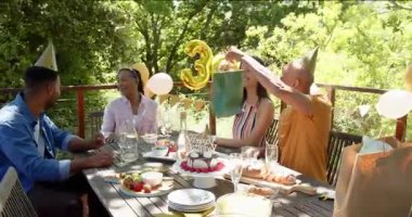 Arkadaşlar güvertede oturuyor, turuncu adam hediye paketini kaldırıyor, kadın hediyesini açıyor, diğerleri alkışlıyor. Hediye, kutlama, bahçe, bahçe, balonlar, pasta, yeşillik.