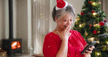 Olgun bir kadın telefon dinlerken gülümsüyor ve yıldız konfetisinin tatil tanıtım mesajına tepki verdiğini gösteriyor. Şenlik, Noel, ağaç, şömine, oturma odası, yetişkin, akıllı telefon