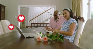 Kadın çiçek düzenlerken erkek arkasına yaslanıyor sosyal medya için dizüstü bilgisayarla kalp yapıyor. Çift, romantik, rahat, ev, yemek, buket, not defteri.