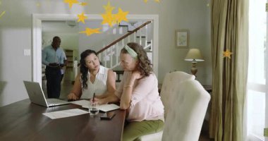 Dizüstü bilgisayarı inceleyen iki kadın, yerleştirme kutusuna erkek olarak giren kağıtlar, ev ekonomisini gösteren altın yıldızlar. Yemek, masa, belgeler, bilgisayar, gün ışığı, işbirliği, konforlu