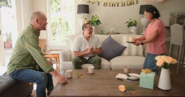 Doğum gününde yanan pastayla gelen kadın, kutlama alkışları, çift kucaklaşması, arkadaş titremesi. Taç, mumlar, bayrak, tatlı, güller, hediye, kanepe