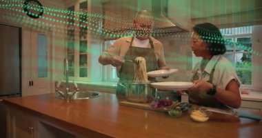 Adam çömlekten makarna kaldırıyor, kadın adada tabak tutuyor, yeşil kaplama yiyecek kaplaması. Çift, mutfak, önlük, servis, maşa, kesme tahtası, ahşap