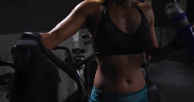 Havlu tutan, spor salonunda bisiklet üstünde mavi şişe tutan, spor sutyen ve şort giyen bir kadın. Kardiyo, fitness, kadın, ter, eldiven, su, siluet