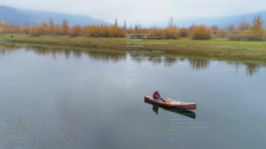Durgun gölde kürek çeken bir adam, turuncu ağaçları kaplayan hareketli bir seyahat haritası çiziyor. Watercraft, solo, paddle, güz, bulut, sükunet, yansımalar
