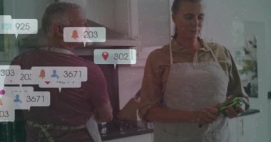 Kıdemli adam esrar karıştırıyor, kadın mutfakta sebze soyuyor sosyal medya baloncukları parıldıyor. Çift, yemek, ev yemeği, soba, önlük, sebze, soyucu.