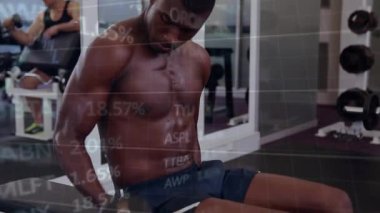 Adam dambılı kavrıyor, güç için kıvrılıyor, borsa tabloları çerçevenin üzerine kayıyor. Sporcu, spor salonu, bank, ağırlık, egzersiz, odak, yoğunluk