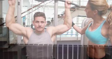 Geniş bir demire tutunan erkek, kadın formunu bulmak için son sürat geri çekiş yapıyor, üstü örtülmüş şekilde. Antrenör, müşteri, takım çalışması, spor salonu, ağırlık, makine, gün ışığı