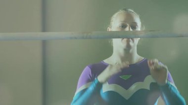 Kadın jimnastikçi avuçlarını ovalıyor sonra yüksek çıtayı kavrıyor, hareketli tebeşir tozu eğitime hazır. Sporcu, aparat, spor salonu, odaklanma, hazırlık, at kuyruğu, deniz mavisi.