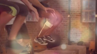 Fitness üçlüsü sıçramaya hazırlanıyor, plyo kutuları üzerinde kutu atlayışı yapıyor, lens alevi altında eğitim gücü sağlıyor. Spor salonu, stüdyo, grup, sporcular, ağırlıklar, tuğla, pencereler