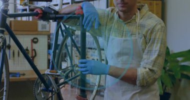 Tamirci bisiklet standını ayarlıyor, kamera geri çekiliyor ve mavi kaplama onarımları gösteriyor. Atölye, bisiklet, eldiven, önlük, teknisyen, bakım, gün ışığı