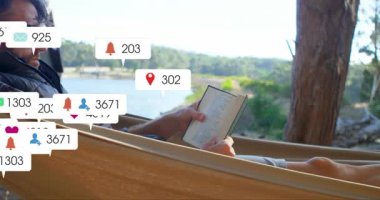 man reclining in hammock by lake reading book, displaying floating social media icons for marketing. Nature, relaxation, outdoor, leisure, tranquility, technology, serenity