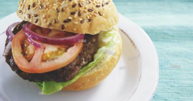 Deri mavisi desenli kumaşın üzerinde beyaz tabakta solda susam tohumu hamburgeri görünüyor. Sandviç, çörek, protein, yeşiller, gevrek, yakın plan, masa üstü.