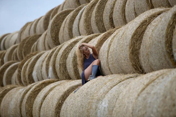 Blonde woman with rubber boots posing in a field on a hay roll