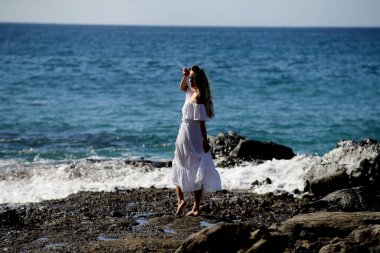 Beyaz giysili çıplak ayaklı bir kız kayalık sahilde yürüyor ve poz veriyor. 