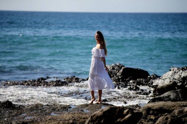 Beyaz giysili çıplak ayaklı bir kız kayalık sahilde yürüyor ve poz veriyor. 