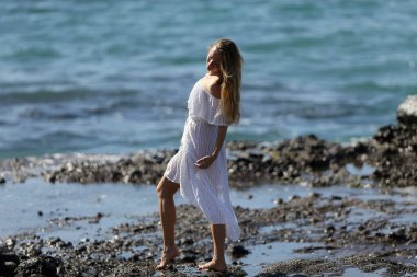 Beyaz giysili çıplak ayaklı bir kız kayalık sahilde yürüyor ve poz veriyor. 