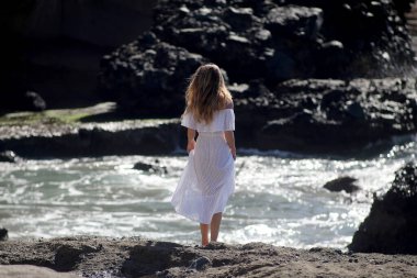 Beyaz giysili çıplak ayaklı bir kız kayalık sahilde yürüyor ve poz veriyor. 