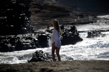 Beyaz giysili çıplak ayaklı bir kız kayalık sahilde yürüyor ve poz veriyor. 