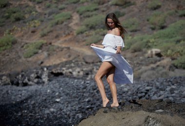 Beyaz giysili çıplak ayaklı bir kız kayalık sahilde yürüyor ve poz veriyor. 