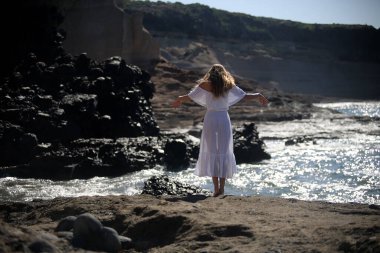 Beyaz giysili çıplak ayaklı bir kız kayalık sahilde yürüyor ve poz veriyor. 