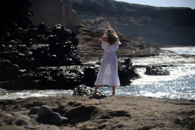 Beyaz giysili çıplak ayaklı bir kız kayalık sahilde yürüyor ve poz veriyor. 