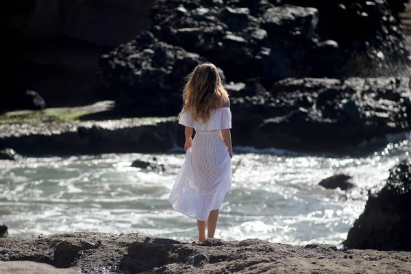 Beyaz giysili çıplak ayaklı bir kız kayalık sahilde yürüyor ve poz veriyor. 