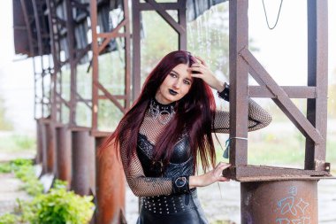 Uzun kızıl saçlı, siyah deri elbiseli ve siyah makyajlı gotik bir manken endüstriyel bir ortamda poz veriyor. Kendine güveni tam bir kadın portresi keskin güzelliğini ve karanlık estetiğini ifade ediyor..