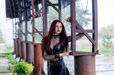 Uzun kızıl saçlı, siyah deri elbiseli ve siyah makyajlı gotik bir manken endüstriyel bir ortamda poz veriyor. Kendine güveni tam bir kadın portresi keskin güzelliğini ve karanlık estetiğini ifade ediyor..