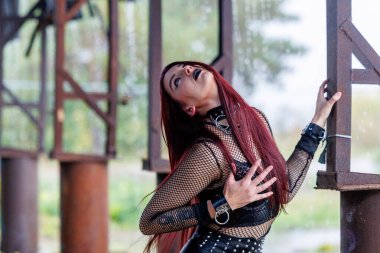 Uzun kızıl saçlı, siyah deri elbiseli ve siyah makyajlı gotik bir manken endüstriyel bir ortamda poz veriyor. Kendine güveni tam bir kadın portresi keskin güzelliğini ve karanlık estetiğini ifade ediyor..