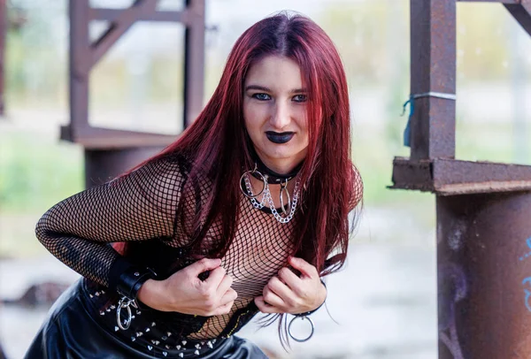 Uzun kızıl saçlı, siyah deri elbiseli ve siyah makyajlı gotik bir manken endüstriyel bir ortamda poz veriyor. Kendine güveni tam bir kadın portresi keskin güzelliğini ve karanlık estetiğini ifade ediyor..