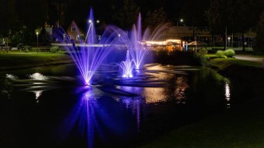 Litvanya 'nın Kazlu Ruda kentindeki sakin bir gölette renkli, aydınlık pınarlar parıldıyor. Yansımalar, park manzarası ve rahat atmosfer seyahat, turizm ve şehir geceleri için mükemmel bir büyülü sahne oluşturur.