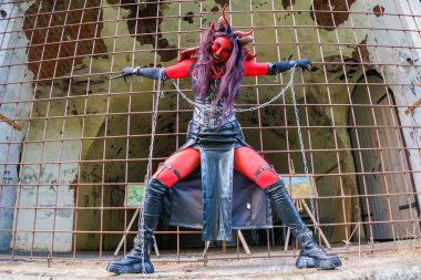 Kırmızı vücut boyalı, boynuzlu ve zincirli şeytani kostümlü bir kadın terk edilmiş zindan ortamında demir parmaklıklar arkasında poz veriyor. Gotik kostümlü karanlık fantezi karakteri ve dramatik makyaj gücü ve yoğunluğu ifade ediyor.