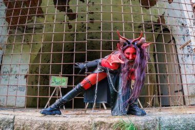Kırmızı vücut boyalı, boynuzlu ve zincirli şeytani kostümlü bir kadın terk edilmiş zindan ortamında demir parmaklıklar arkasında poz veriyor. Gotik kostümlü karanlık fantezi karakteri ve dramatik makyaj gücü ve yoğunluğu ifade ediyor.