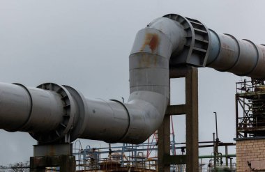 Bulutlu bir günde beton sütunlar tarafından desteklenen büyük endüstriyel boru hattı. Metal yüzey, çıplak ağaçlar ve sessiz manzaralarla çevrili yıpranma ve pas belirtileri gösteriyor.