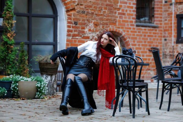 Стильная молодая женщина в красном шарфе и черных высоких сапогах сидит в открытом городском кафе. Осенняя атмосфера, элегантный стиль одежды и уверенное присутствие. Редакционное использование
