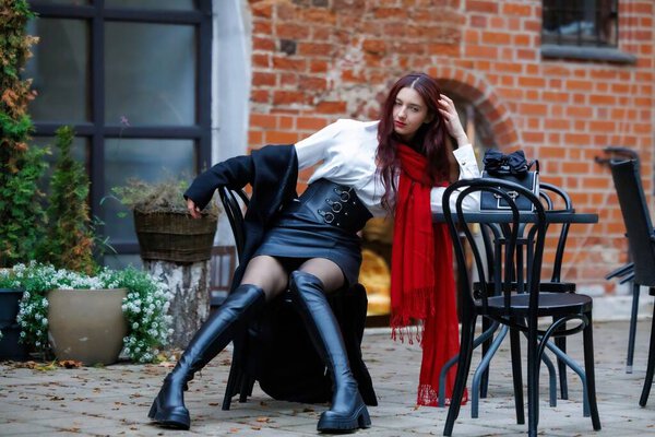 Стильная молодая женщина в красном шарфе и черных высоких сапогах сидит в открытом городском кафе. Осенняя атмосфера, элегантный стиль одежды и уверенное присутствие. Редакционное использование