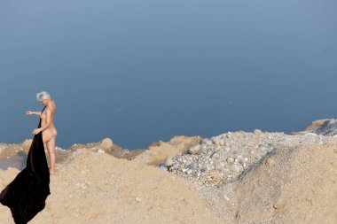 Doğada Zarafet: Karanlık Kumaşlı Bir Kadın Silueti Bir Arkaplan Su ve Kuma Karşı