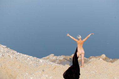 Doğada Zarafet: Karanlık Kumaşlı Bir Kadın Silueti Bir Arkaplan Su ve Kuma Karşı