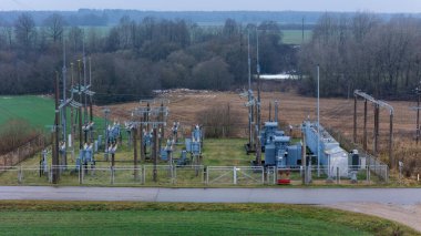 Elektrik hatları, trafolar, açık tarım alanları ve bulutlu gökyüzünün altındaki uzak ormanlarla çevrili bir kırsal elektrik santrali doğal kırsal kesimde endüstriyel varlığı ifade ediyor.