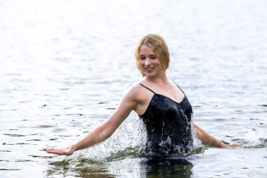 Siyah elbisesi dalgalanırken sakin suda zarif dans hareketleri yapan etkileyici genç bir kadın. Özgürlük kavramı, kadınlık, denge, duygusal ifade, beden farkındalığı ve doğa ile uyum.