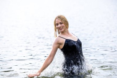 Siyah elbisesi dalgalanırken sakin suda zarif dans hareketleri yapan etkileyici genç bir kadın. Özgürlük kavramı, kadınlık, denge, duygusal ifade, beden farkındalığı ve doğa ile uyum.