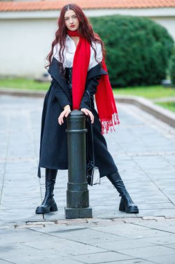 Tarihi bir kasaba ortamında canlı bir sonbahar portresi. Uzun siyah bir ceket ve kırmızı eşarp giyen genç bir kadın metal bir direğin yanında güvenle duruyor. Yumuşak doğal ışık, sığ alan derinliği ve ılık mevsimsel ruh hali sanatsal bir belgesel portresi oluşturur.