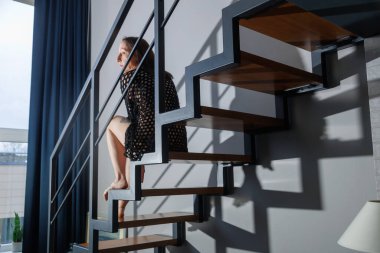 Modern merdivenlerde oturan kadın, yumuşak doğal ışık altında. Minimalist iç mekan, geometrik çizgiler ve gölgeler, sakin ruh hali, çağdaş yaşam tarzı, mahremiyet, yalnızlık, modern ev mimarisi