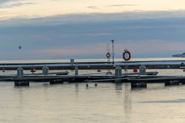 Nida Limanı 'nda kışın boş bir marina. Buzla kaplı yüzen iskeleler, sakin sular, bulutlu gökyüzü ve kıyı manzarası. İnsan yok, tekne yok. Soğuk mevsim, sessizlik, asgari deniz geçmişi.