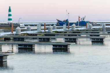Nida Limanı 'nda kışın boş bir marina. Buzla kaplı yüzen iskeleler, sakin sular, bulutlu gökyüzü ve kıyı manzarası. İnsan yok, tekne yok. Soğuk mevsim, sessizlik, asgari deniz geçmişi.