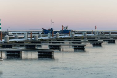 Nida Limanı 'nda kışın boş bir marina. Buzla kaplı yüzen iskeleler, sakin sular, bulutlu gökyüzü ve kıyı manzarası. İnsan yok, tekne yok. Soğuk mevsim, sessizlik, asgari deniz geçmişi.