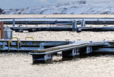Nida Limanı 'nda kışın boş bir marina. Buzla kaplı yüzen iskeleler, sakin sular, bulutlu gökyüzü ve kıyı manzarası. İnsan yok, tekne yok. Soğuk mevsim, sessizlik, asgari deniz geçmişi.