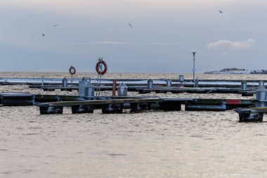 Nida Limanı 'nda kışın boş bir marina. Buzla kaplı yüzen iskeleler, sakin sular, bulutlu gökyüzü ve kıyı manzarası. İnsan yok, tekne yok. Soğuk mevsim, sessizlik, asgari deniz geçmişi.