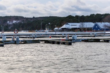 Nida Limanı 'nda kışın boş bir marina. Buzla kaplı yüzen iskeleler, sakin sular, bulutlu gökyüzü ve kıyı manzarası. İnsan yok, tekne yok. Soğuk mevsim, sessizlik, asgari deniz geçmişi.