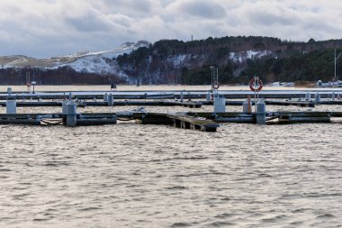 Nida Limanı 'nda kışın boş bir marina. Buzla kaplı yüzen iskeleler, sakin sular, bulutlu gökyüzü ve kıyı manzarası. İnsan yok, tekne yok. Soğuk mevsim, sessizlik, asgari deniz geçmişi.